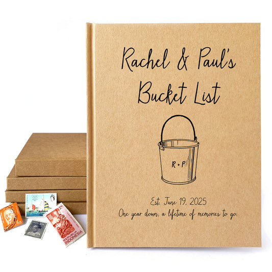 Personalized bucket list book for couples standing next to a stack of handbound books and old stamps. Cover is personalized with their names and a favorite quote.