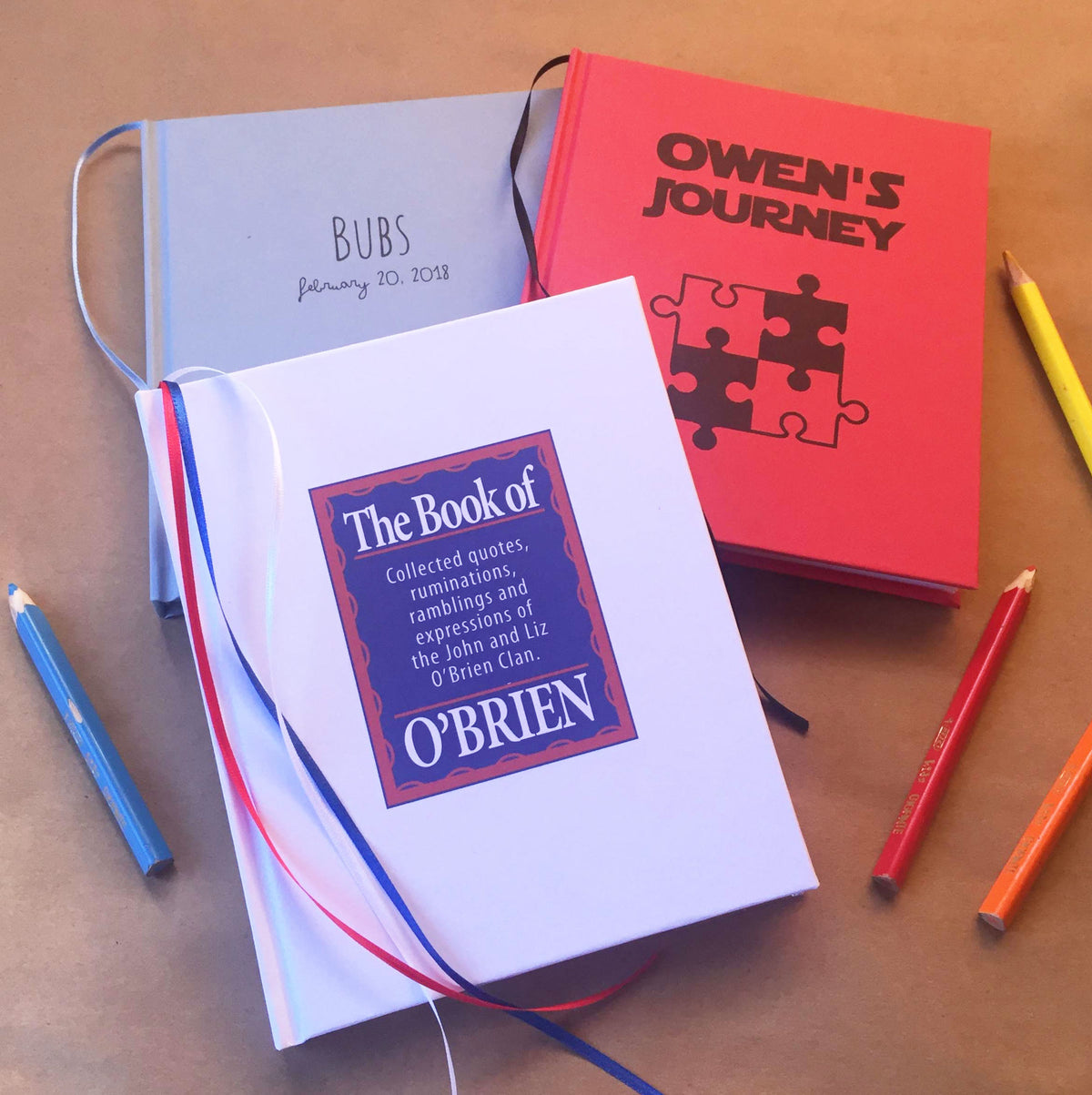 3 custom journal covers in gray, red and white. Each custom printed with it's own text and design. On a beige background next to colored pencils.