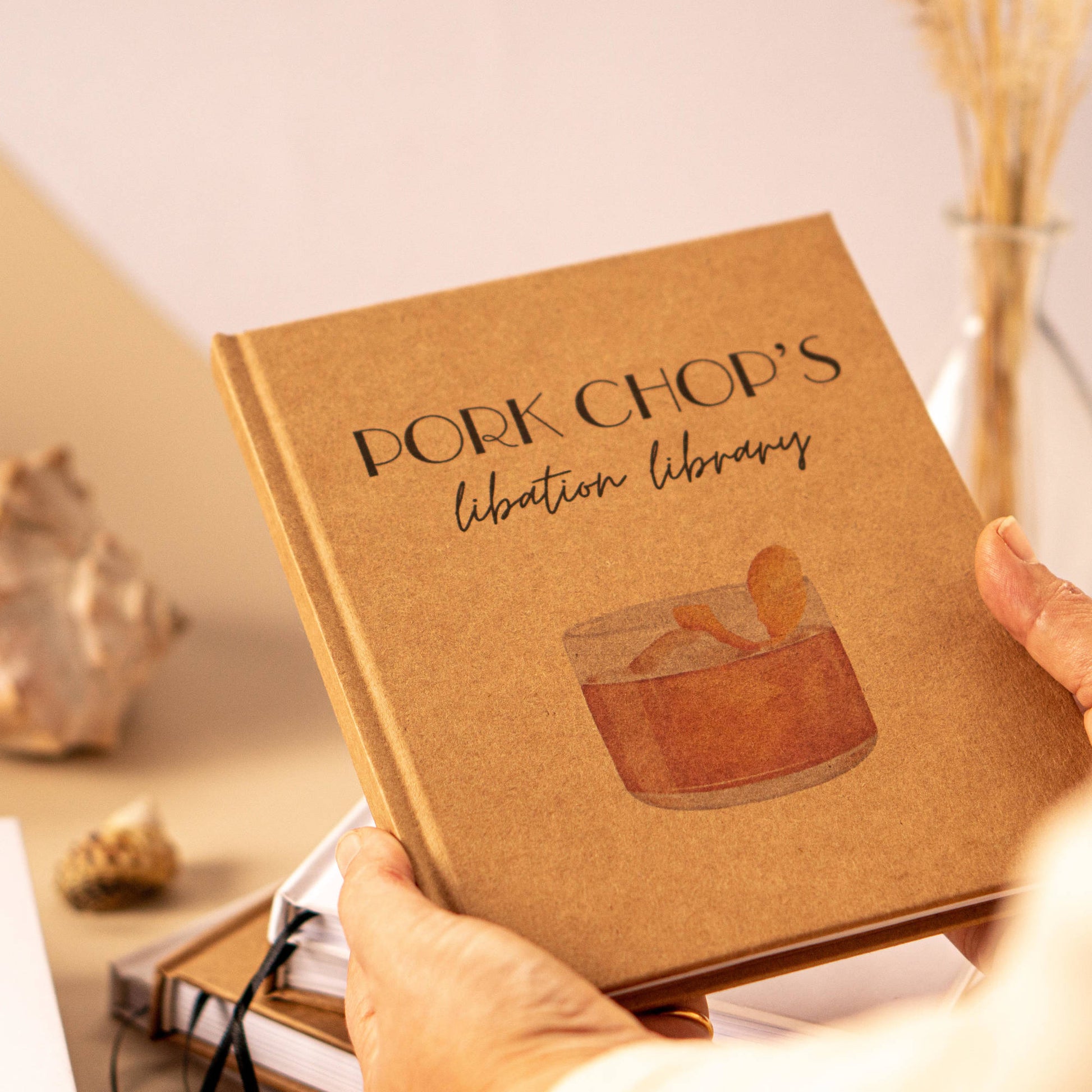 Two hands holding a personalized cocktail book with custom text and drink illustration on cover. Handcrafted book, heirloom quality.