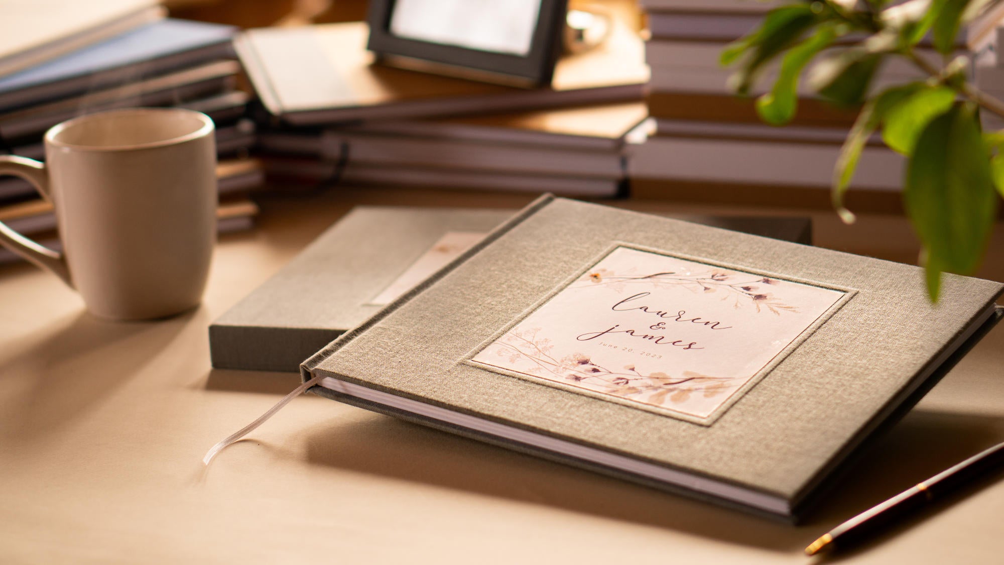 Personalized wedding guest book with gray linen cover. Hardback with personalized flower bouquet cover design with custom names and date. On table with pen, mug and other customized books.