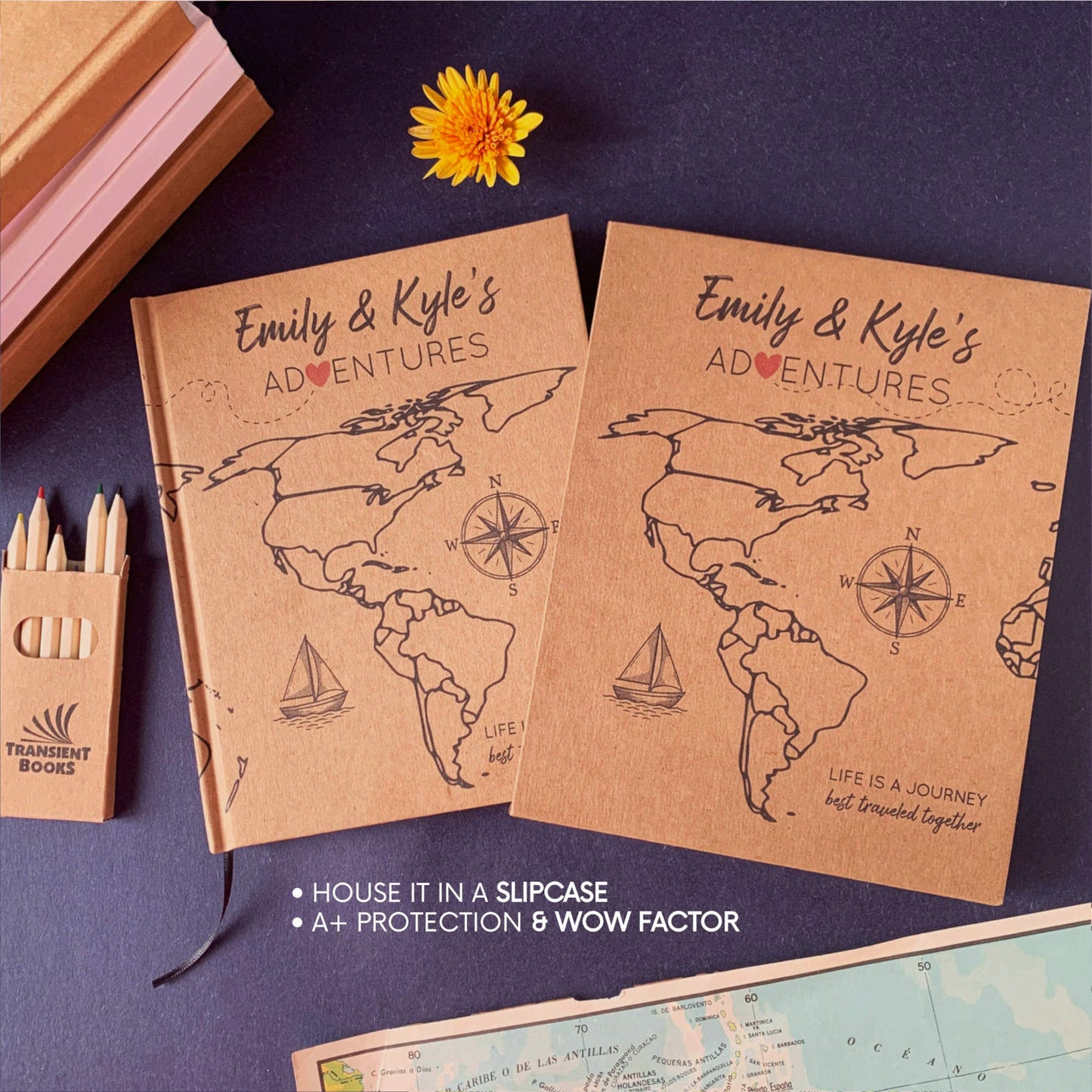 A personalized travel journal with world map design with a matching book slipcase on a navy blue surface with a yellow flower. Colored pencils and map on the table. Book is handbound, hardback.