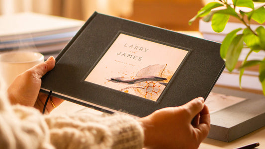A personalized wedding guest book with branded wedding artwork on the cover being held by two hands.