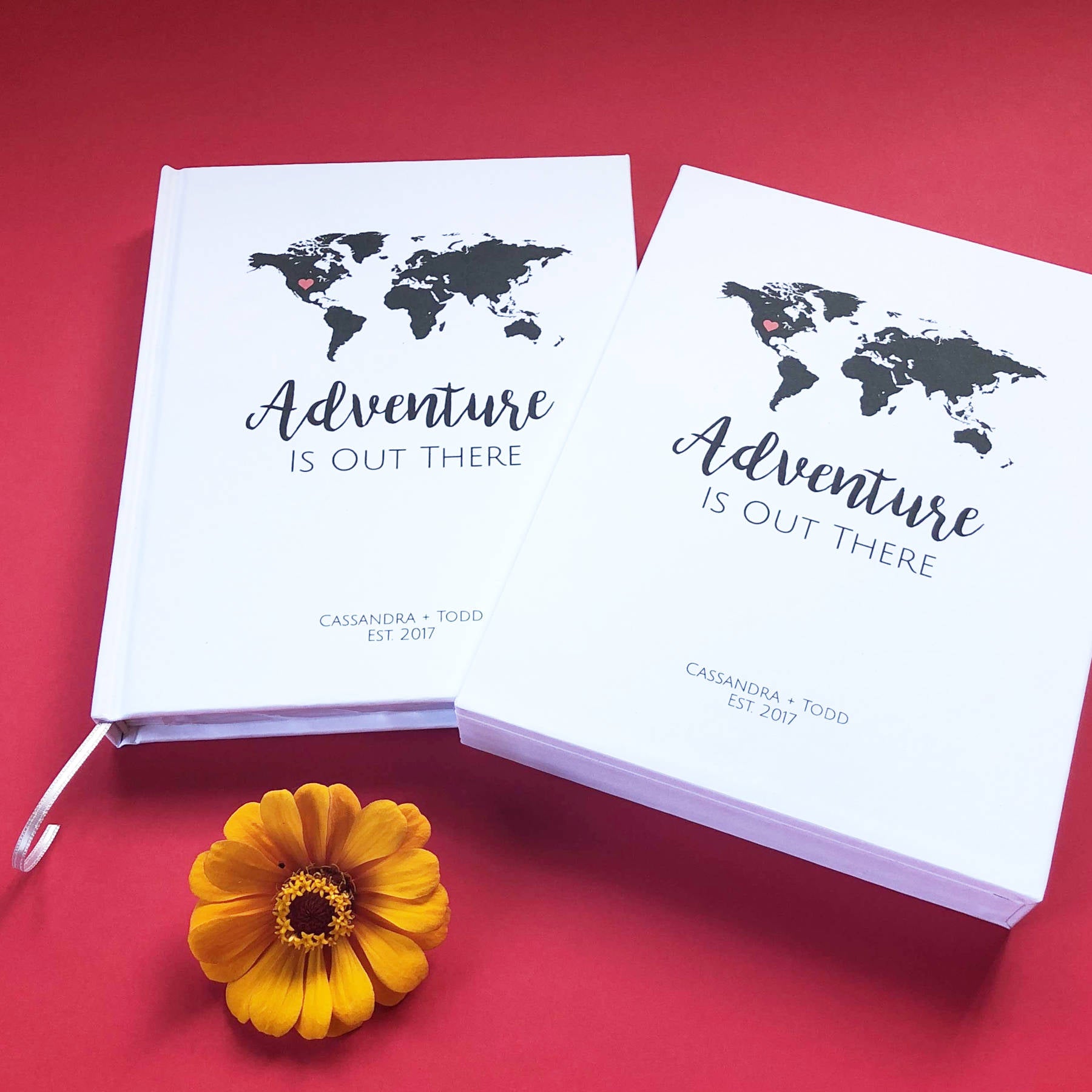 White adventure journal for couples with world map design and custom text on the front, housed in a matching book slipcase on a red table.
