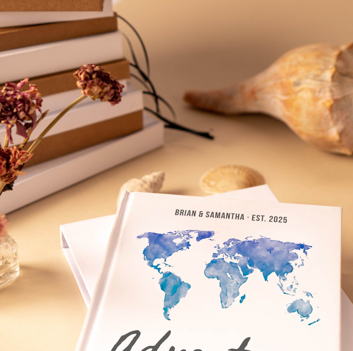 Adventure journal for couples with a blue water color world map design and custom text on a table next to a stack of blank journals, dried flowers and sea shells.