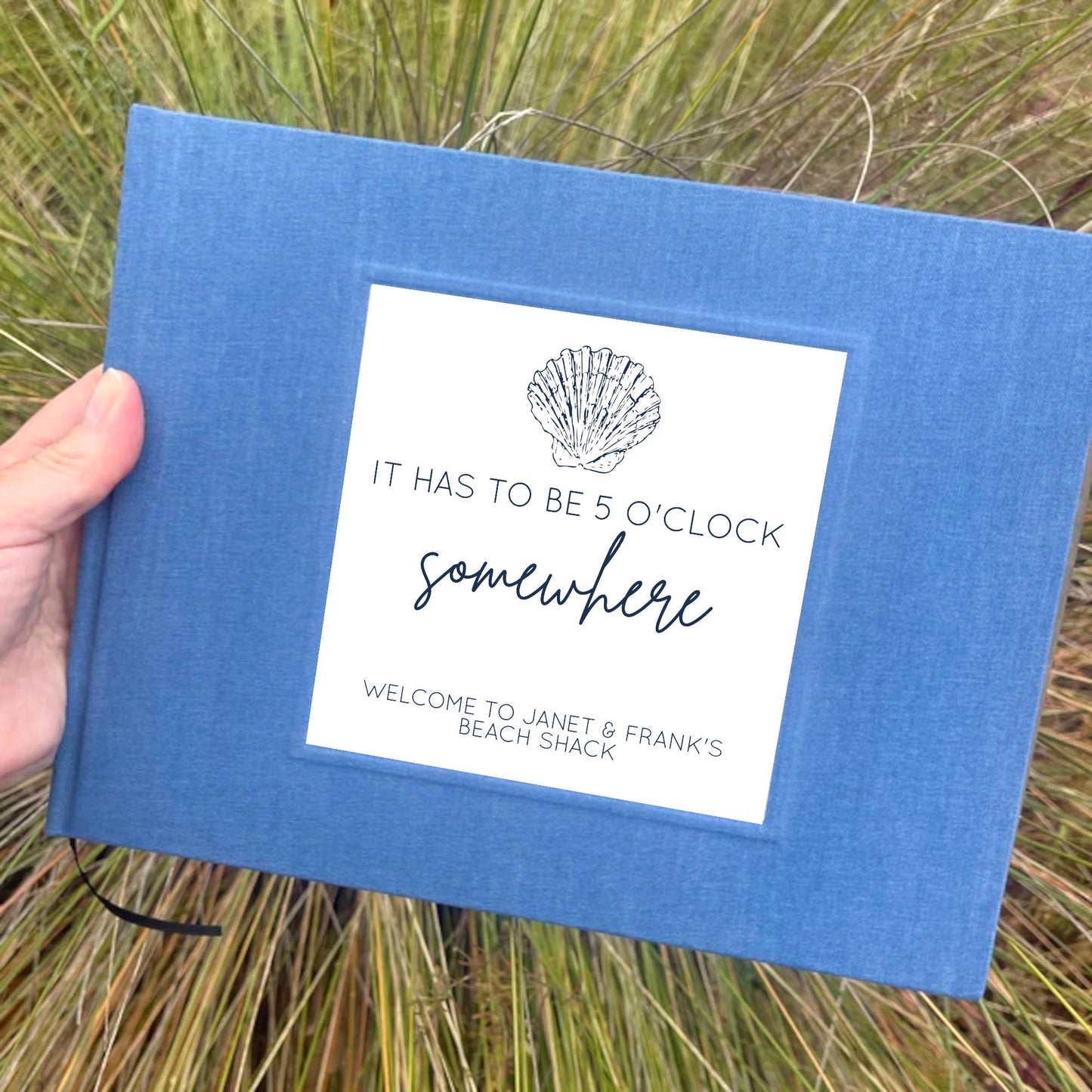 Hand holding a blue beach house guest book in front of wild grasses. Hard back, heirloom quality.