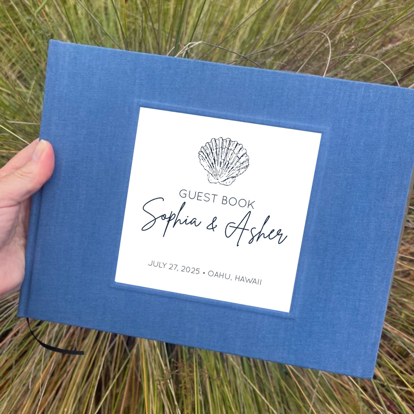 Navy blue linen beach wedding guest book with personalized names on the cover in front of a beach grass background.