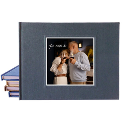 A dark slate gray hardcover guest book shown closed, with a printed sample photo insert on the front and a stack of similar albums fanned out on the left against a white background.