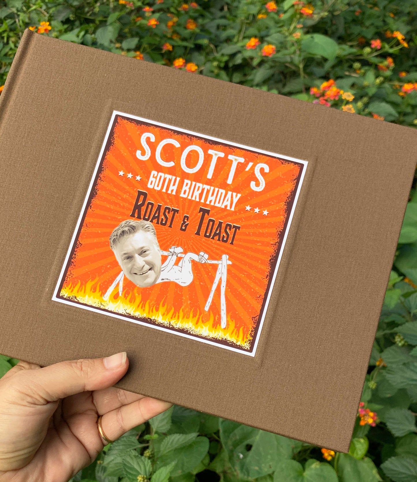 Brown linen 60th birthday party guest book with a custom cover featuring 'Scott's 60th Birthday Roast & Toast' design held by a hand against a green background with flowers.