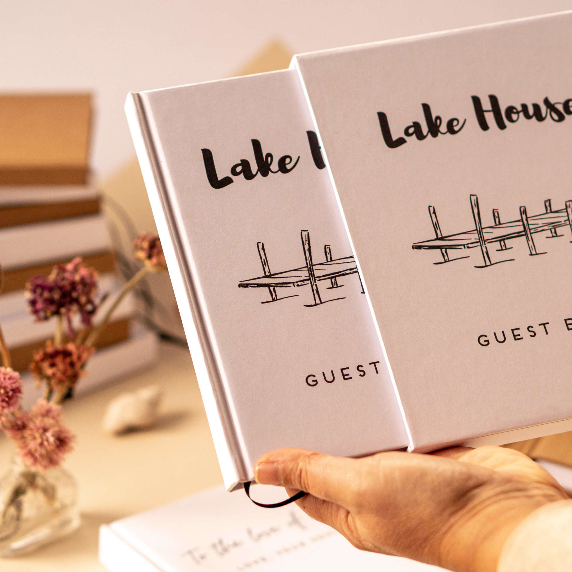 Personalized hand bound lake house guest book housed in a matching book slip case for protection. Both are being hand held in front of a table displaying custom handmade books.