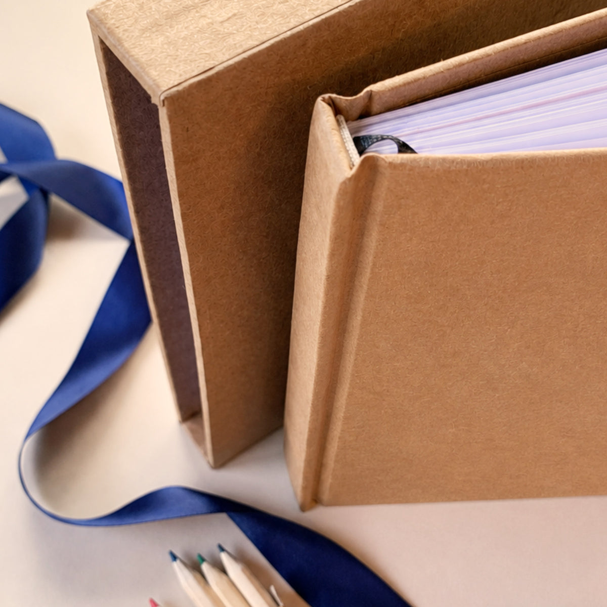 A close-up of a brown kraft paper book slipcase standing open, with a blue ribbon draped across the surface and a few colored pencils at the bottom. No person, animal, or real environment is visible, so this is Product/Packaging rather than Lifestyle.