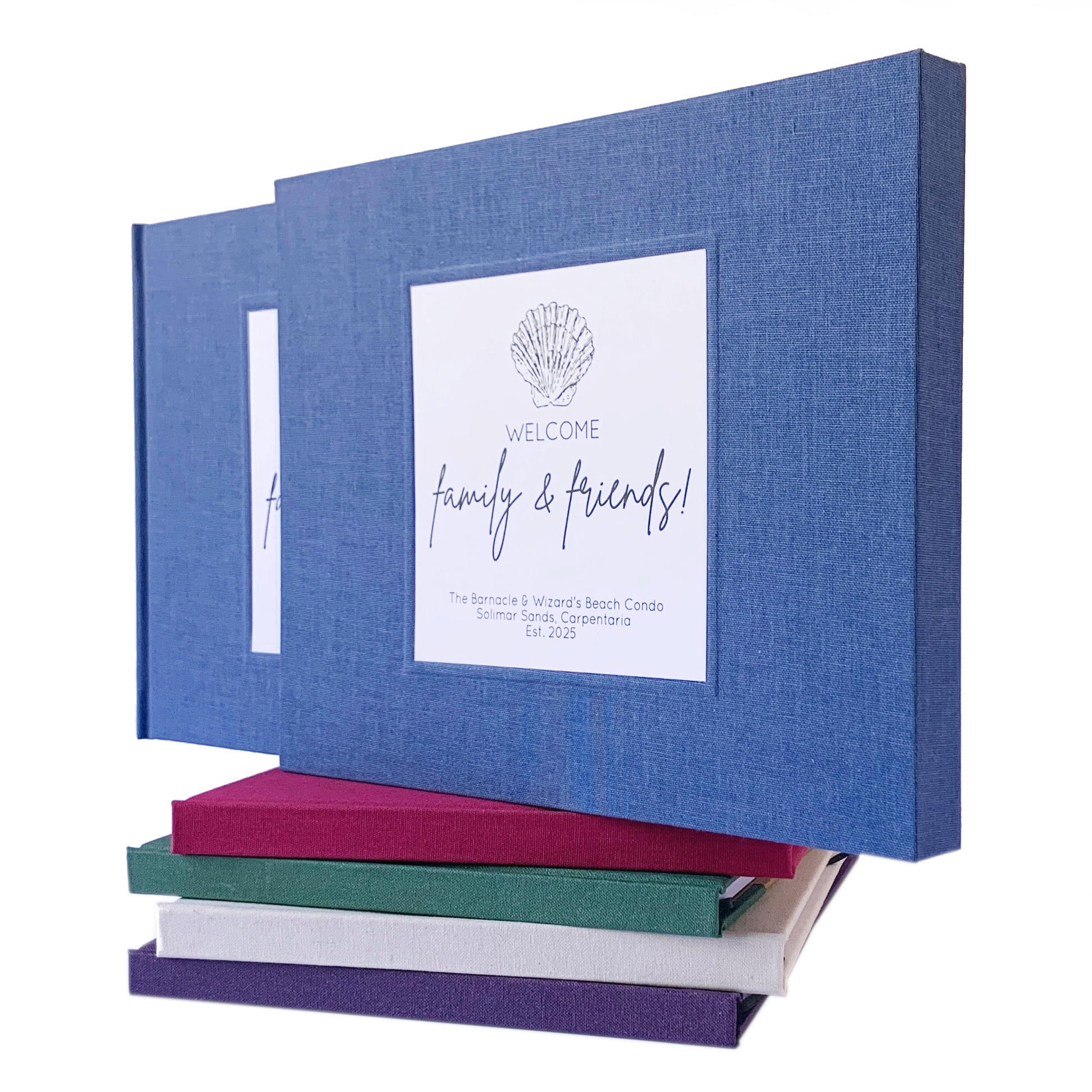 Blue linen beach house guest book in a matching book slipcase. Personalized artwork on front with a coastal theme. Book is on top of a stack of multi colored handbound guest books.