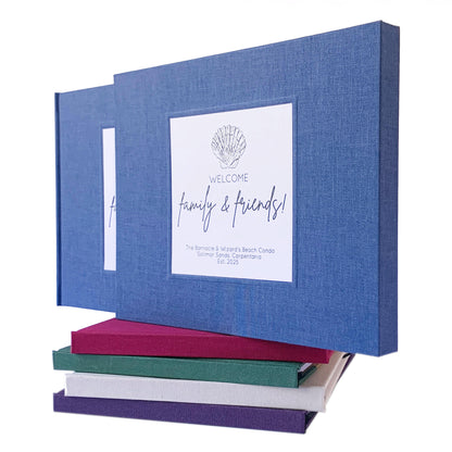 Blue linen beach house guest book in a matching book slipcase. Personalized artwork on front with a coastal theme. Book is on top of a stack of multi colored handbound guest books.