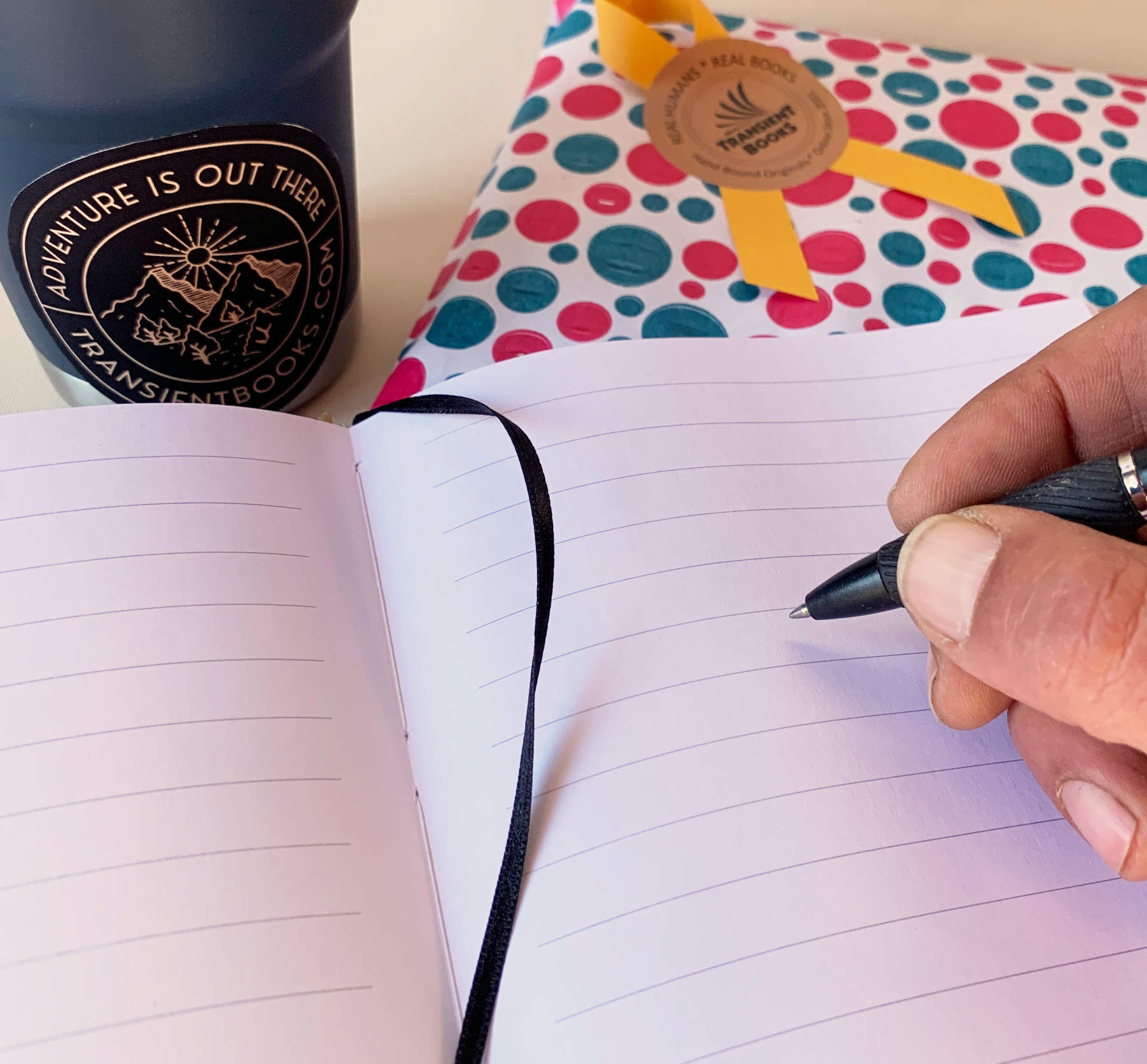 Person writing in a layflat notebook with lined pages with a mug and polka dot giftwrap in the background