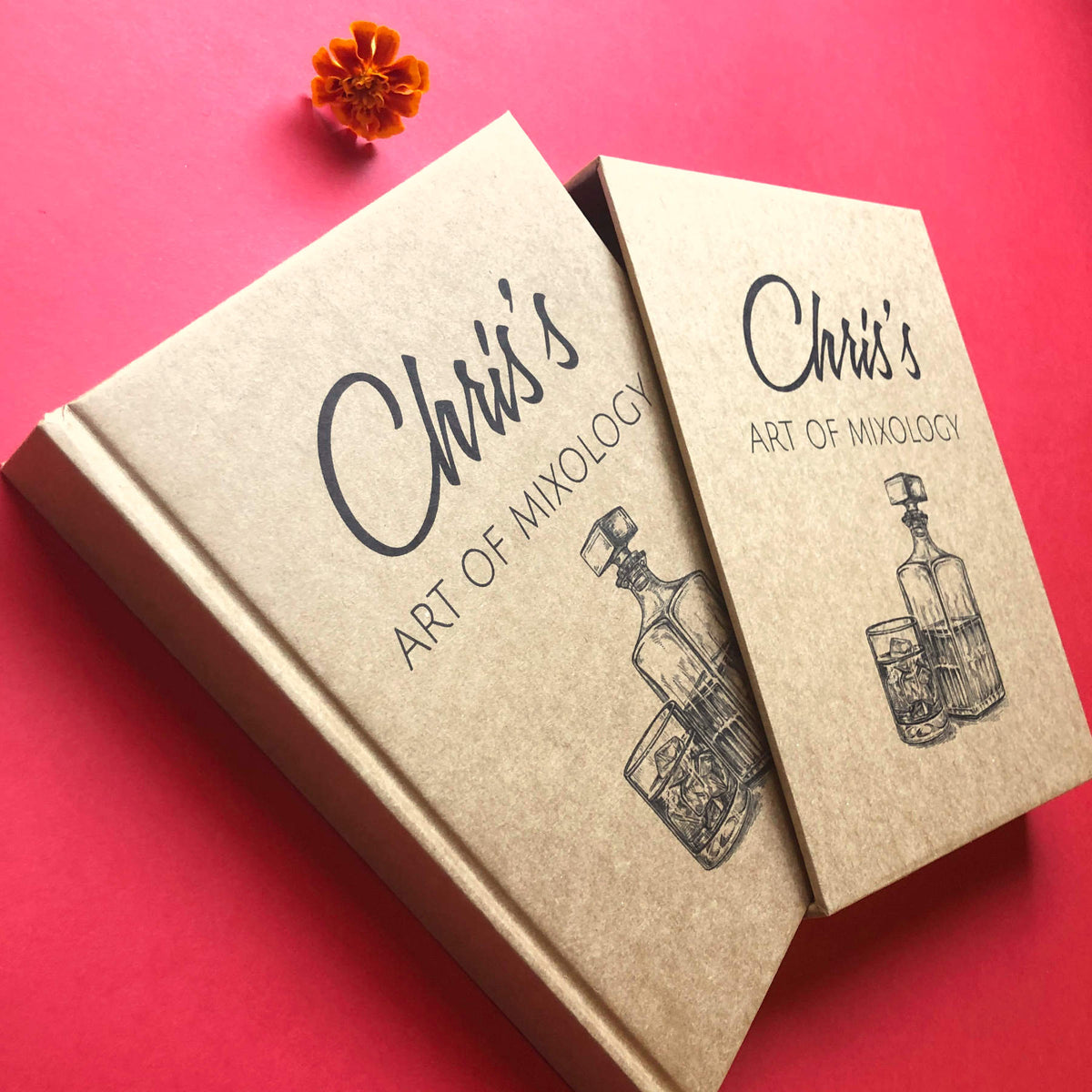 Personalized cocktail recipe book with a kraft cover, custom text, and an illustration of a decanter and a whiskey glass. Housed in a matching book slipcase. On a red table.
