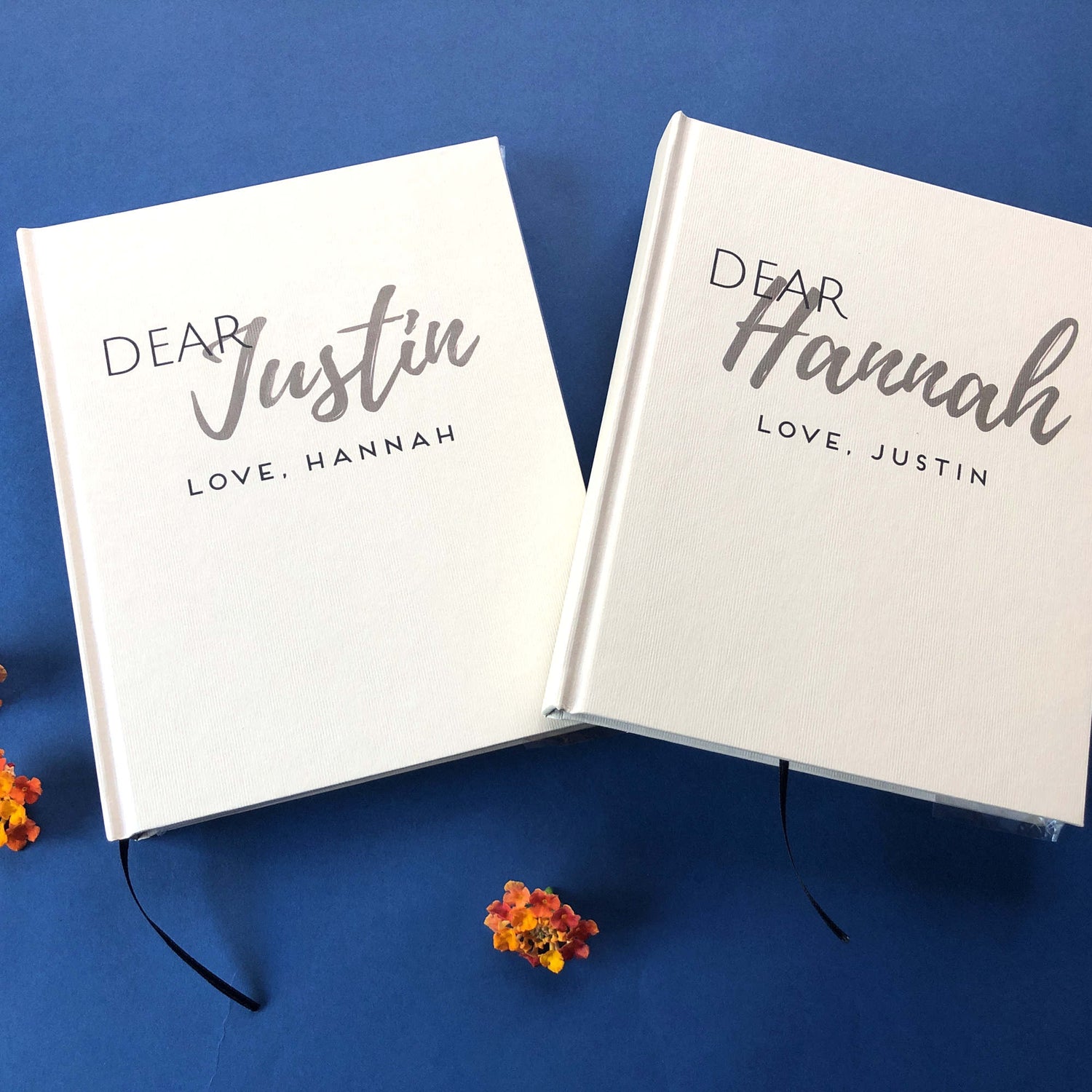 A matching set of couples journals that are letters to husband from wife, letters to wife from husband with custom names on hardback covers. Modern, understated design, hand crafted books on a navy blue surface with orange flowers.