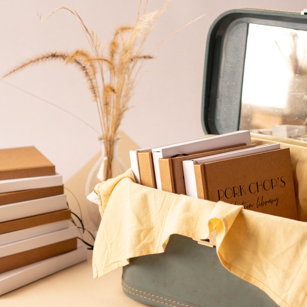 A stack of kraft colored and white custom blank journals on a desk next to a vase of dried grass, next to a vintage blue suitcase full of more custom and kraft colored handbound journals.