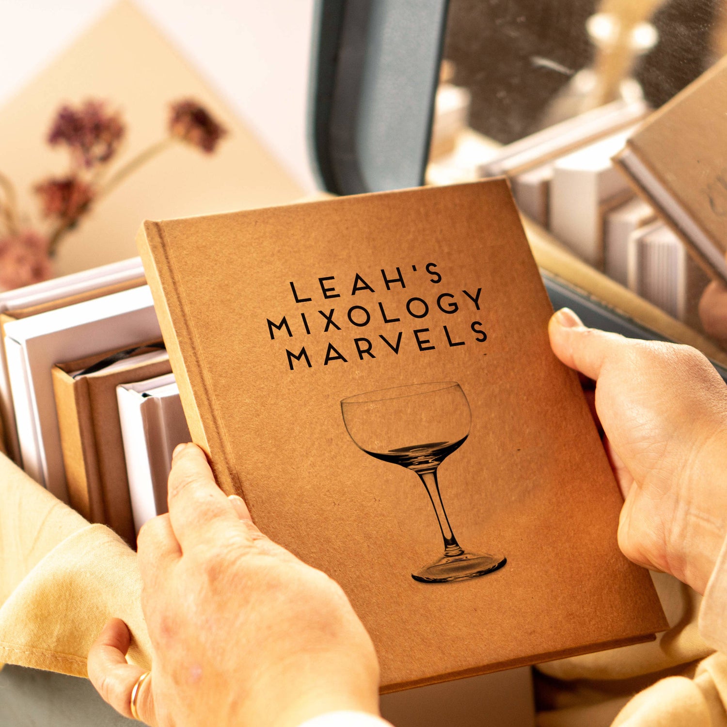 A custom cocktail book being held by two hands. The cover is personalized with the text and a coupe glass.