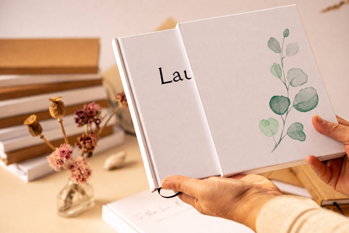 White hardback journal, custm printed with leaves and text, housed in a book slipcase, being handheld in front of dried flowers and a stack of books.