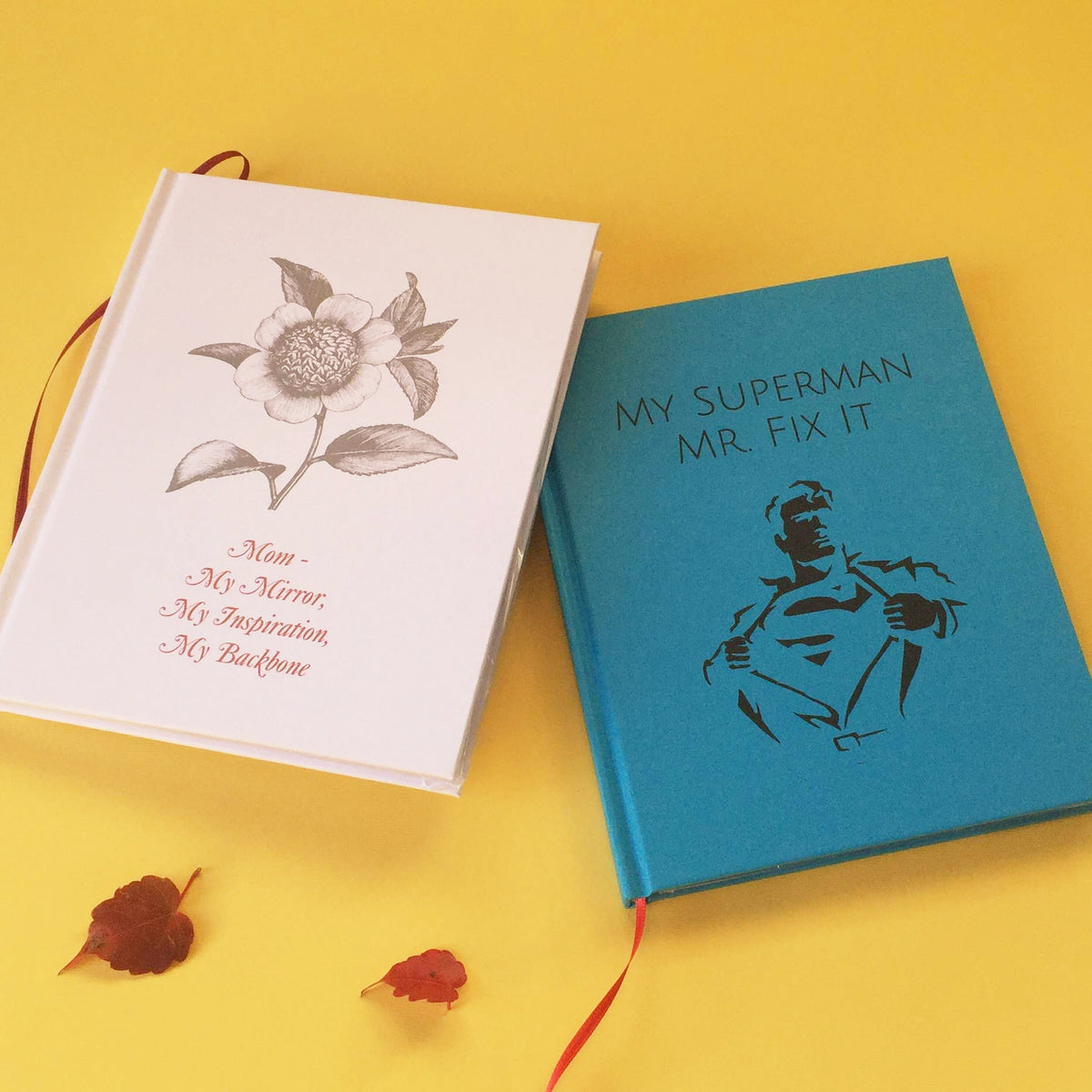 A white custom journal with a flowers and a dedication to a mother next to a turquoise custom journal with superman and a funny superman title on the cover on a yellow background next to 2 red leaves.