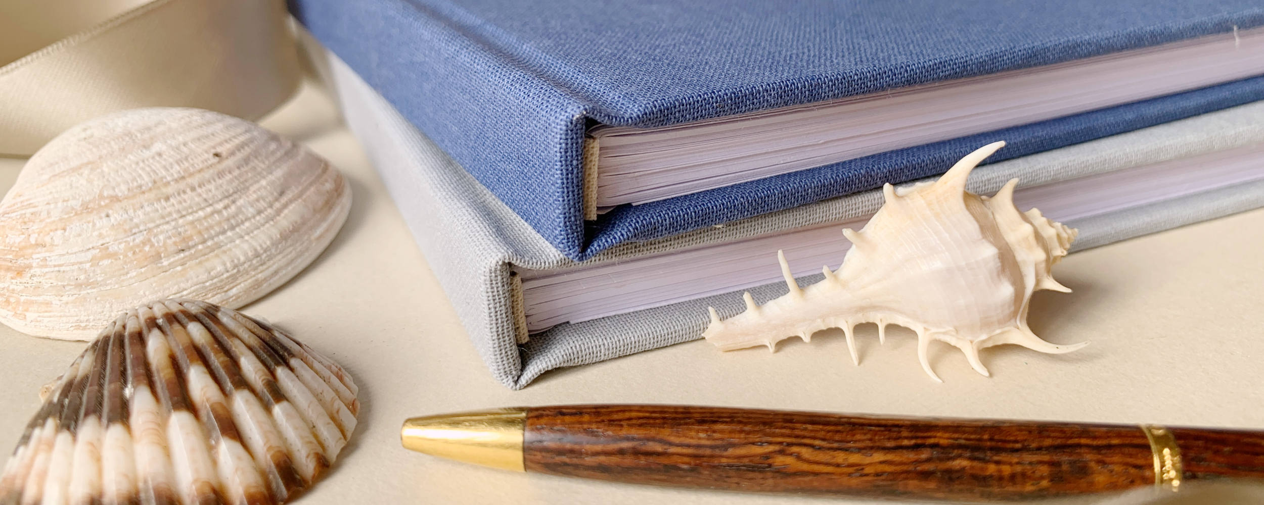 Blue and gray linen guest books on a table with sea shells and a pen.