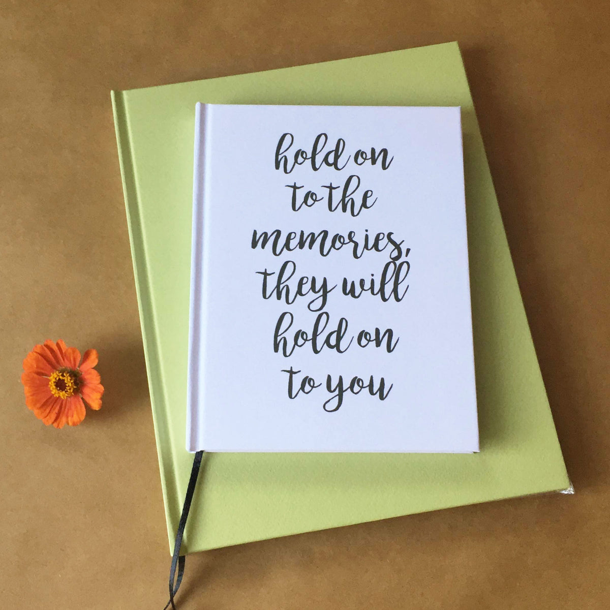A white custom printed journal with the quote "hold onto the memories or they will hold onto you" on the cover, sitting on top of a bright green blank journal, on a beige background next to an orange flower.