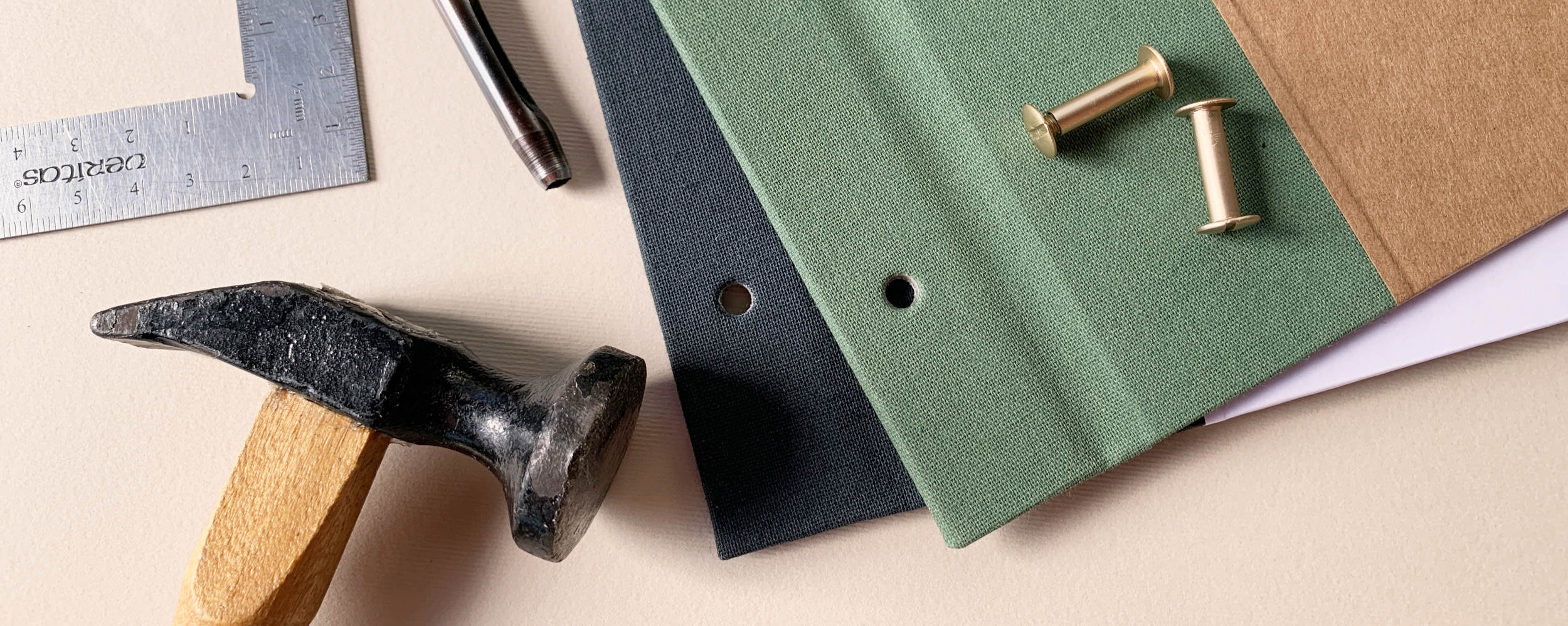 Close up of custom brass screwpost scrapbook albums with hammer and hole punch used to make holes for posts, next to a metal ruler, on tabletop.