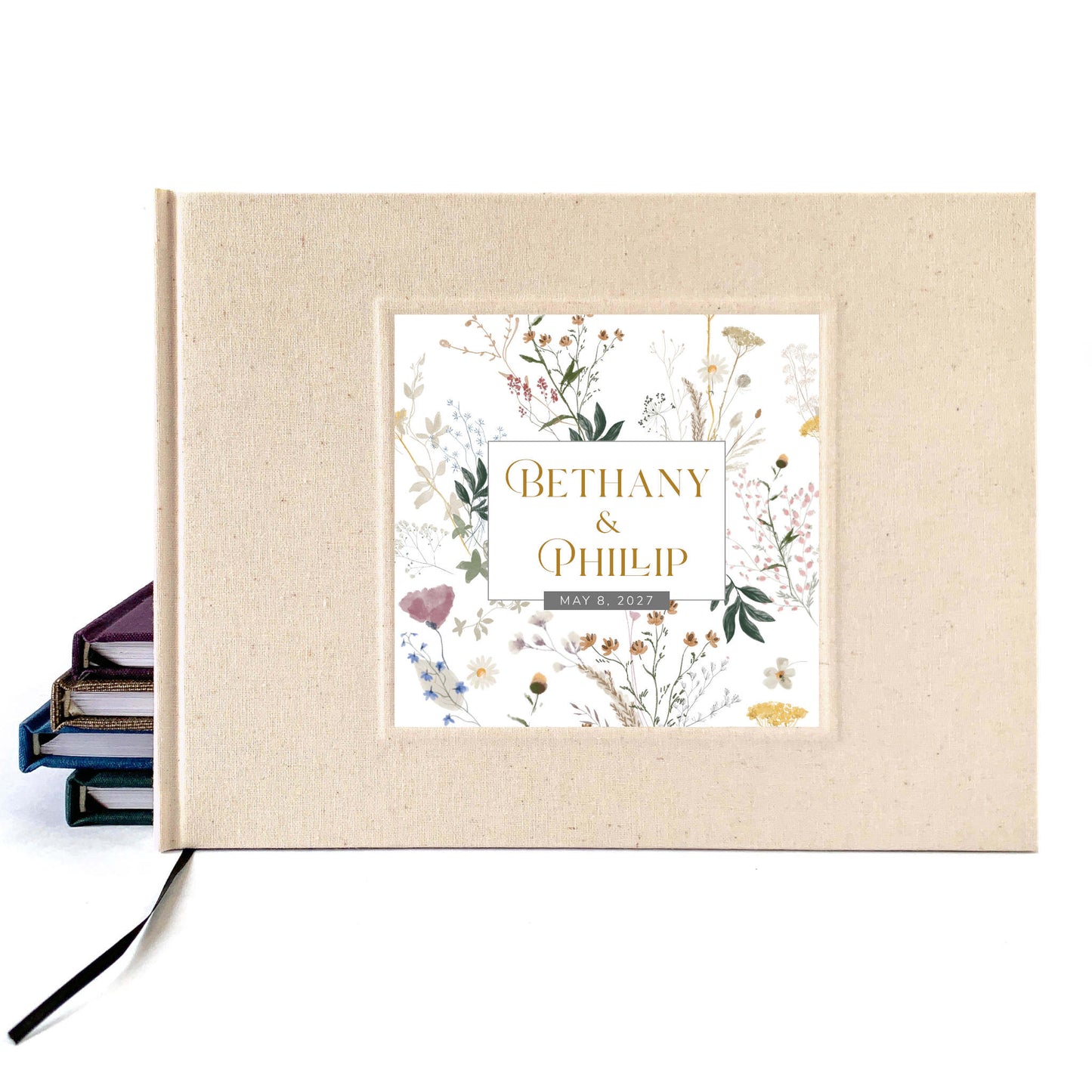 A personalized hardcover wedding guest book with a cream linen cover and a central garden floral illustration panel featuring the names 'Bethany & Phillip' in gold lettering. A black ribbon bookmark is visible on the left, resting against a white background.
