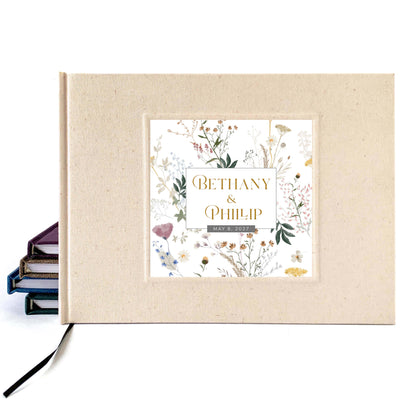 A personalized hardcover wedding guest book with a cream linen cover and a central garden floral illustration panel featuring the names 'Bethany & Phillip' in gold lettering. A black ribbon bookmark is visible on the left, resting against a white background.