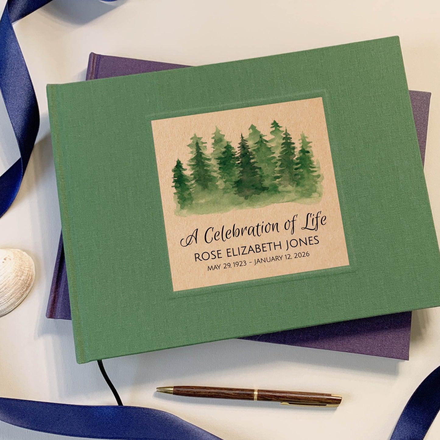 Green funeral guest book on a table with pens, shells and a ribbon with personalized watercolor forest artwork and text.
