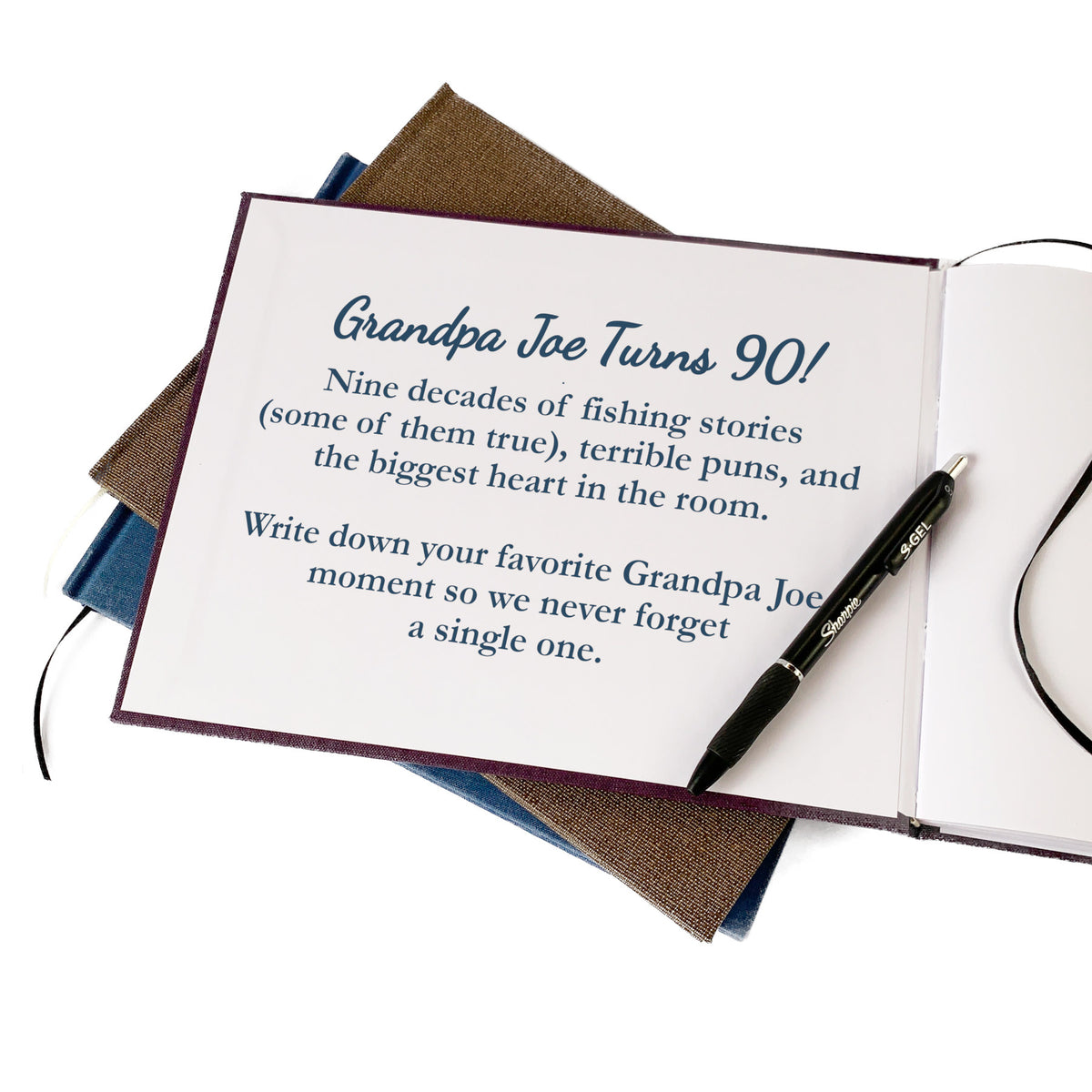 A hardback guest book idea for a 90th birthday party, open to the inside front cover that has a meaningful dedication for Grandpa Joe. A pen lays on the open page. The open book is stacked on top of two more books. The background is white.