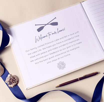 Open vacation home guest book with a welcome message and two oars on a beige surface with a pen and shell.