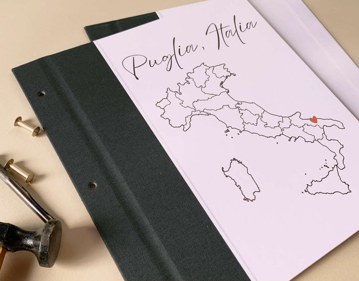 Travel scrapbook notebook with a travel map of Puglia, Italy on a beige background with brass screwposts and a hammer.
