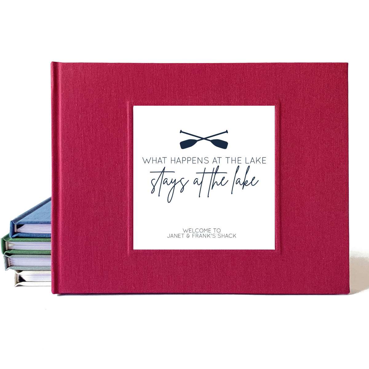 Burgundy linen custom guest book for lake house airbnb vacation rental with white cover inlay personalized with the family's name, crossed oars and "What happens at the lake, stays at the lake" on a white backdrop leaning on a set of books.