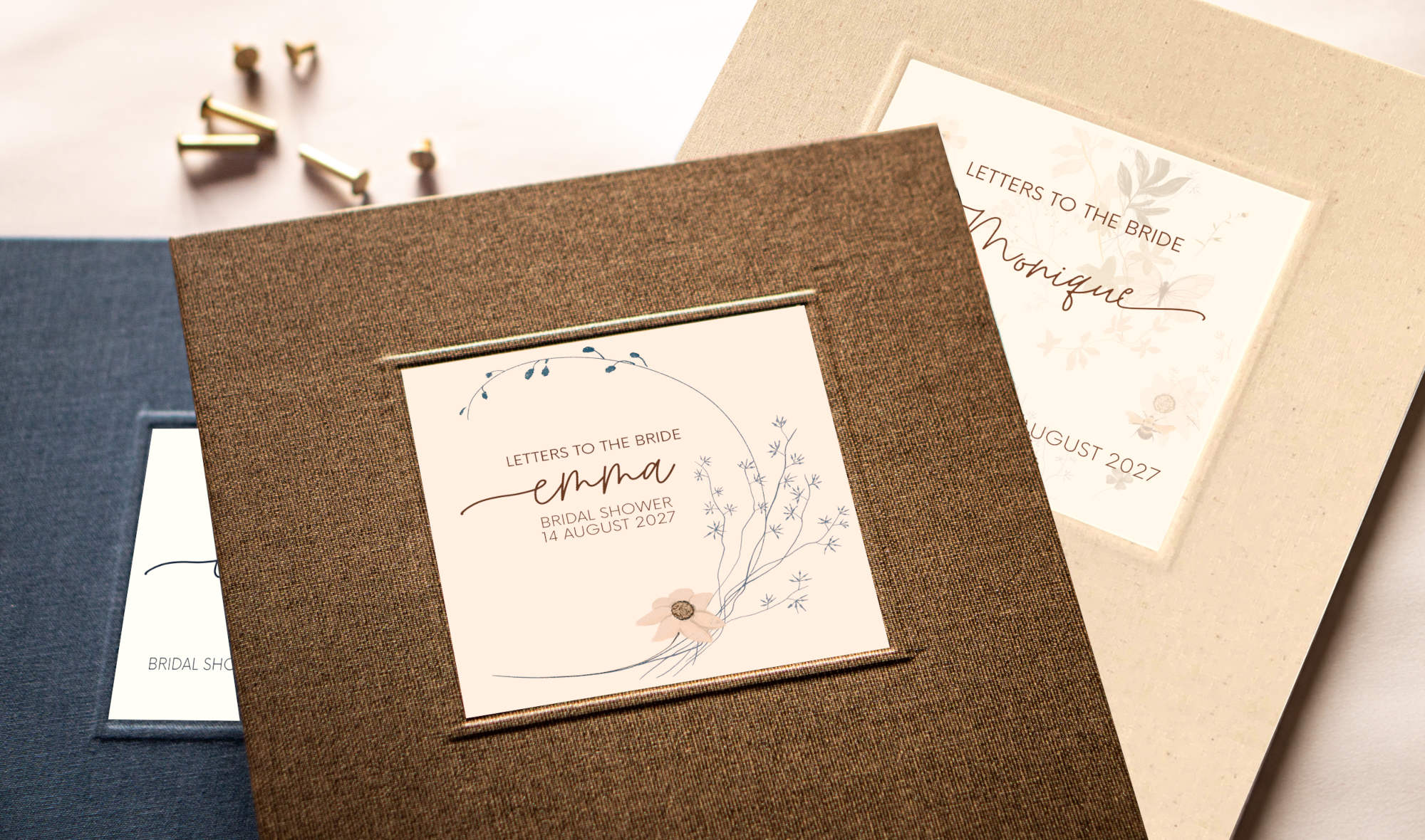 Three personalized "Letters to the Bride" books in blue, brown and cream linen with custom printed cover inlays laying on a table next to brass screw posts.