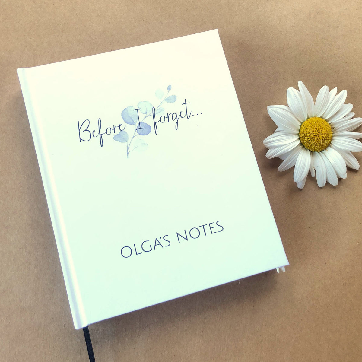 A personalized white journal with the title "Before I forget..." above the words "Olga's Notes" on a beige background next to a daisy.