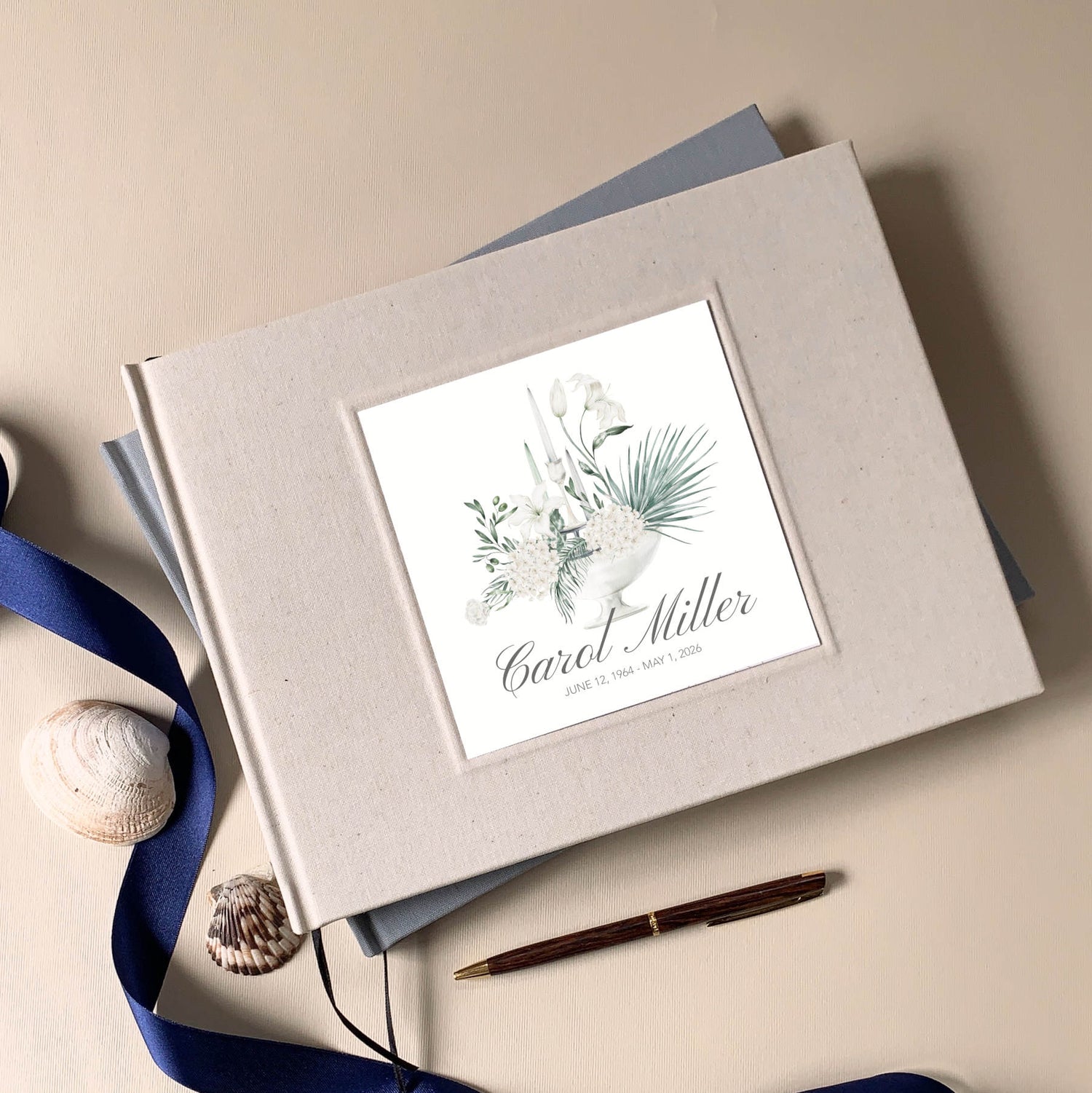 Personalized memorial guest book with white floral bouquet design and custom name and dates on the cover, on a memory table with pen and sea shells.