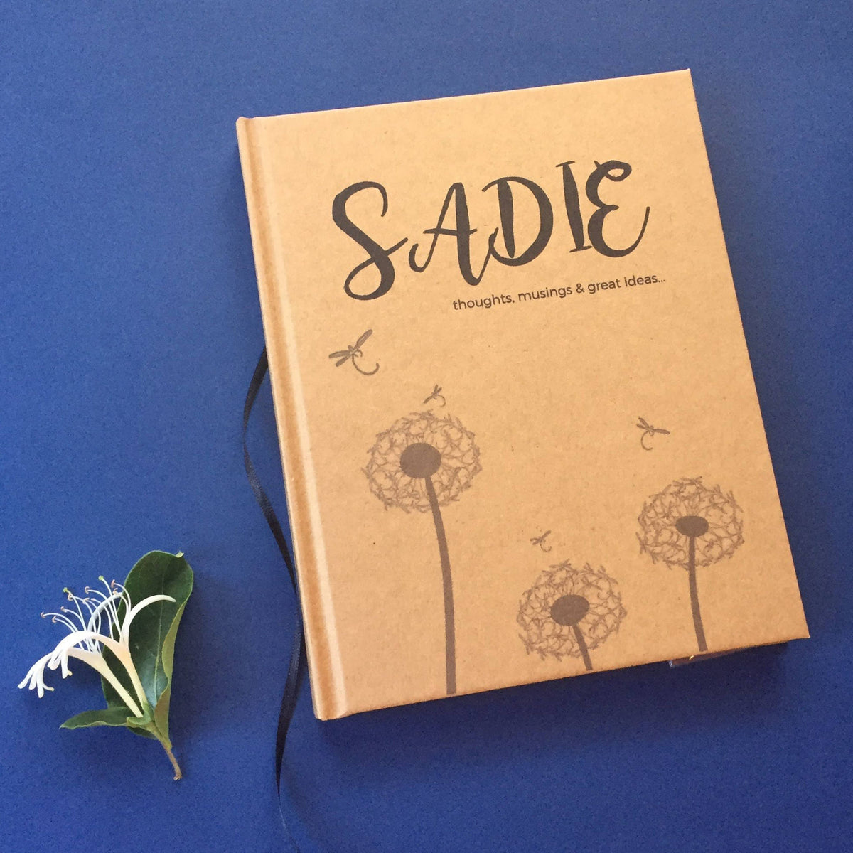 A kraft colored name journal with the name Sadie at the top above a dandelion and dragonfly graphic with the words "thoughts, musings & great ideas..." on a navy blue back ground next to a honeysuckle flower.