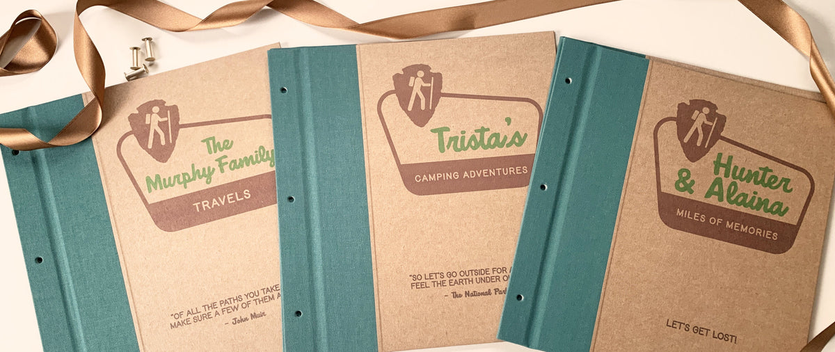 Three National Parks Scrapbooks on a beige table with brass screwposts and brown ribbon. Their covers have personalized text on national parks looking signs with inspirational travel quotes below. The covers have forest green spine canvas and kraft cover paper. The holes are at the 3 ring binder location. 