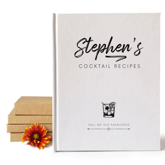 White custom cocktail book with personalized text and old fashioned drink art on the cover, standing next to a stack of handbound books and orange flower.