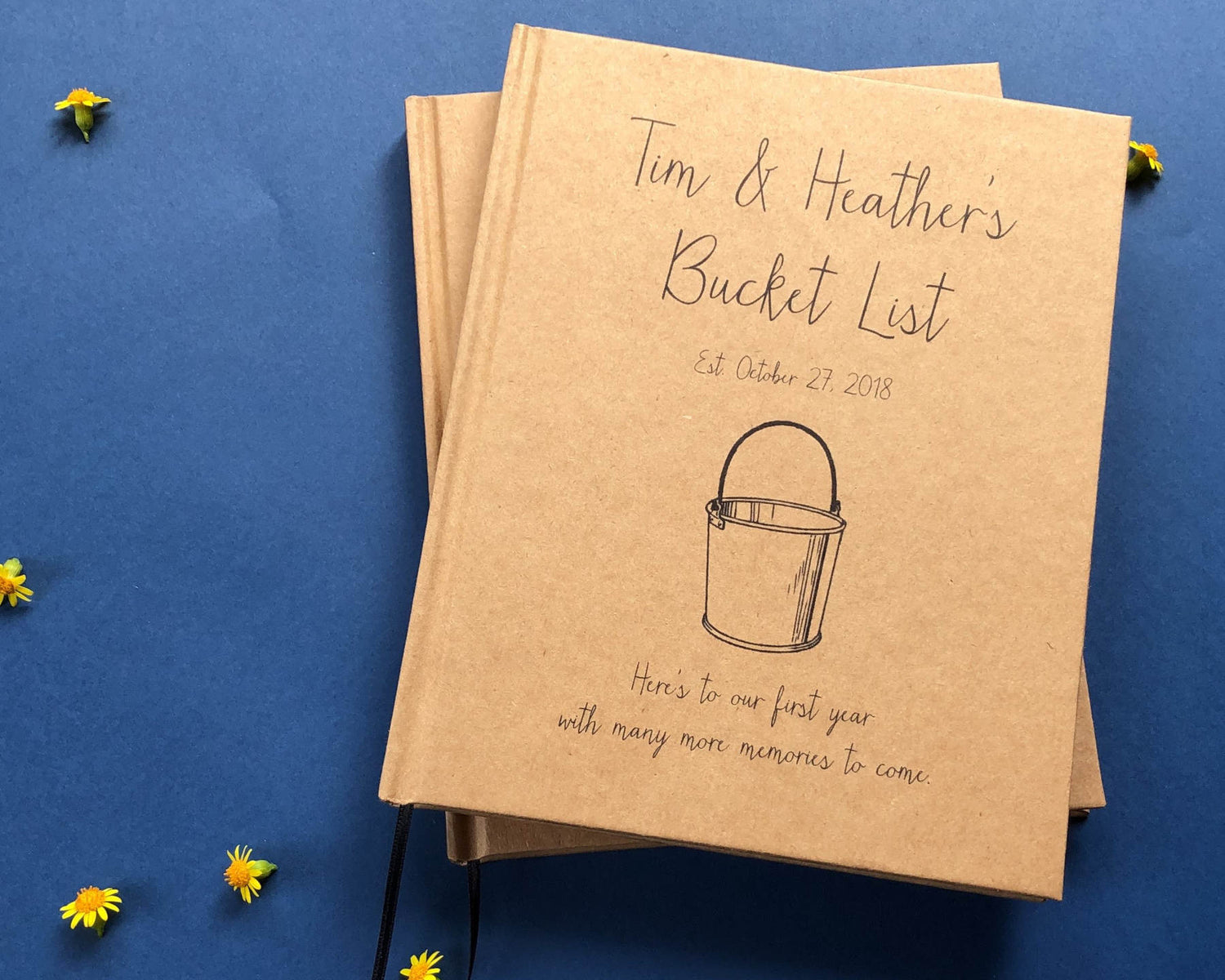 A kraft covered personalized couple's bucket list book with custom text and a bucket drawing on the cover, on a blue table with little yellow flowers.