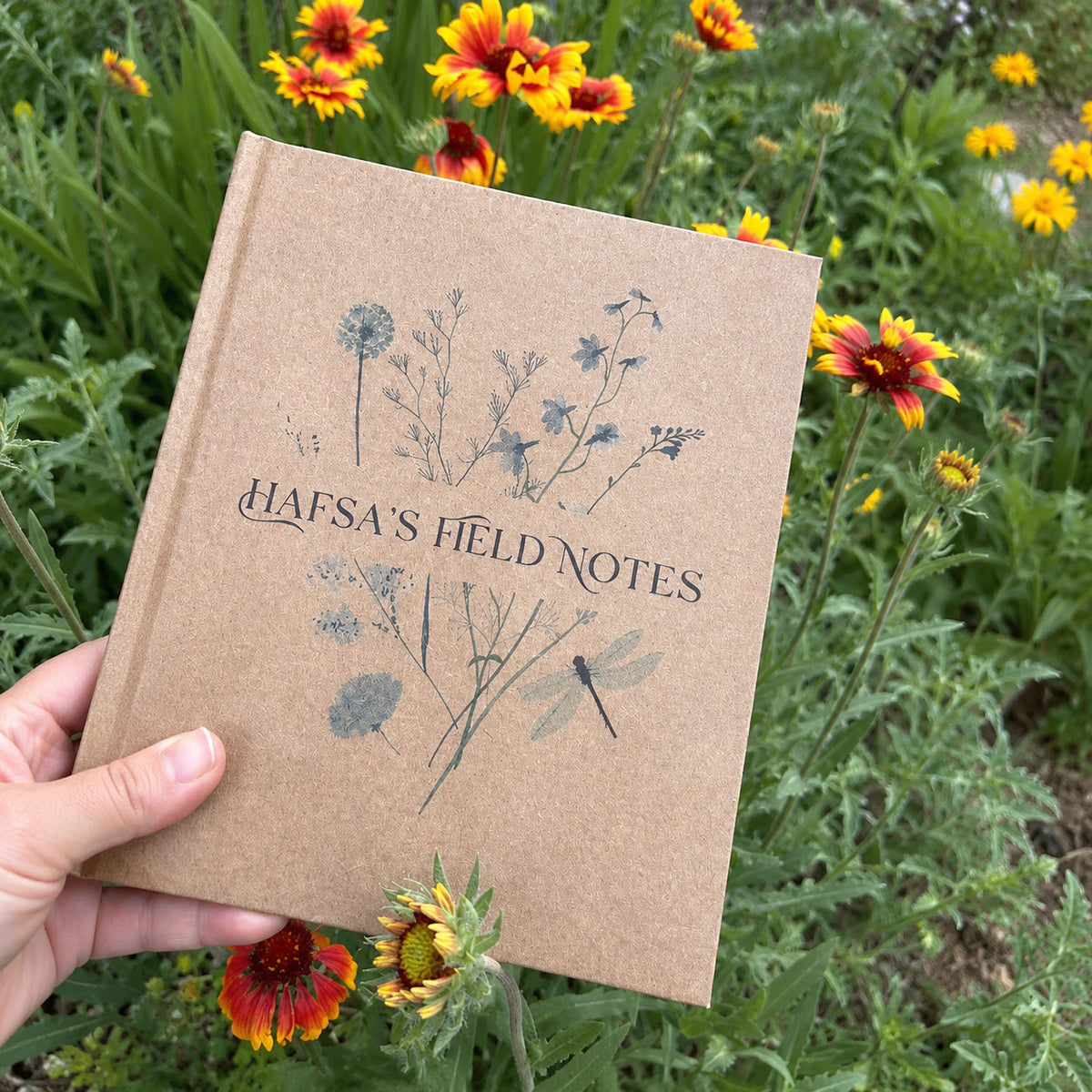 Personalized garden dairy entitled "Hafsa's Field Notes" with a blue bouquet design on the cover with a dragon fly held by a hand in front of a very green flower bed full of yellow and orange flowers.