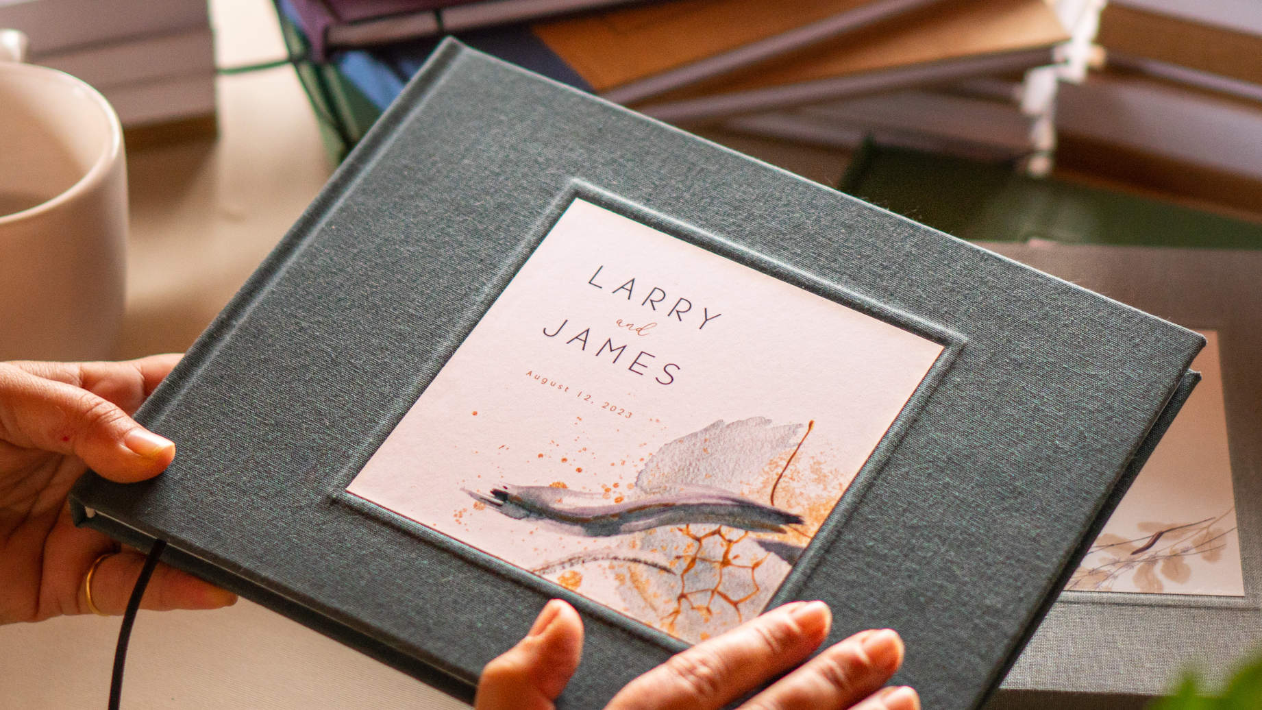 Personalized wedding guest book with gray linen cover. Hardback with personalized flower bouquet cover design with custom names and date. On table with mug and other customized books.
