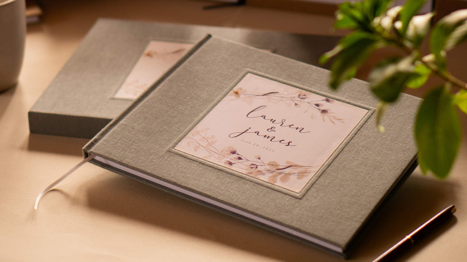 Custom wedding guest book with gray linen cover. Hardback with personalized flower bouquet cover design with custom names and date. On table with pen, mug and other customized books.