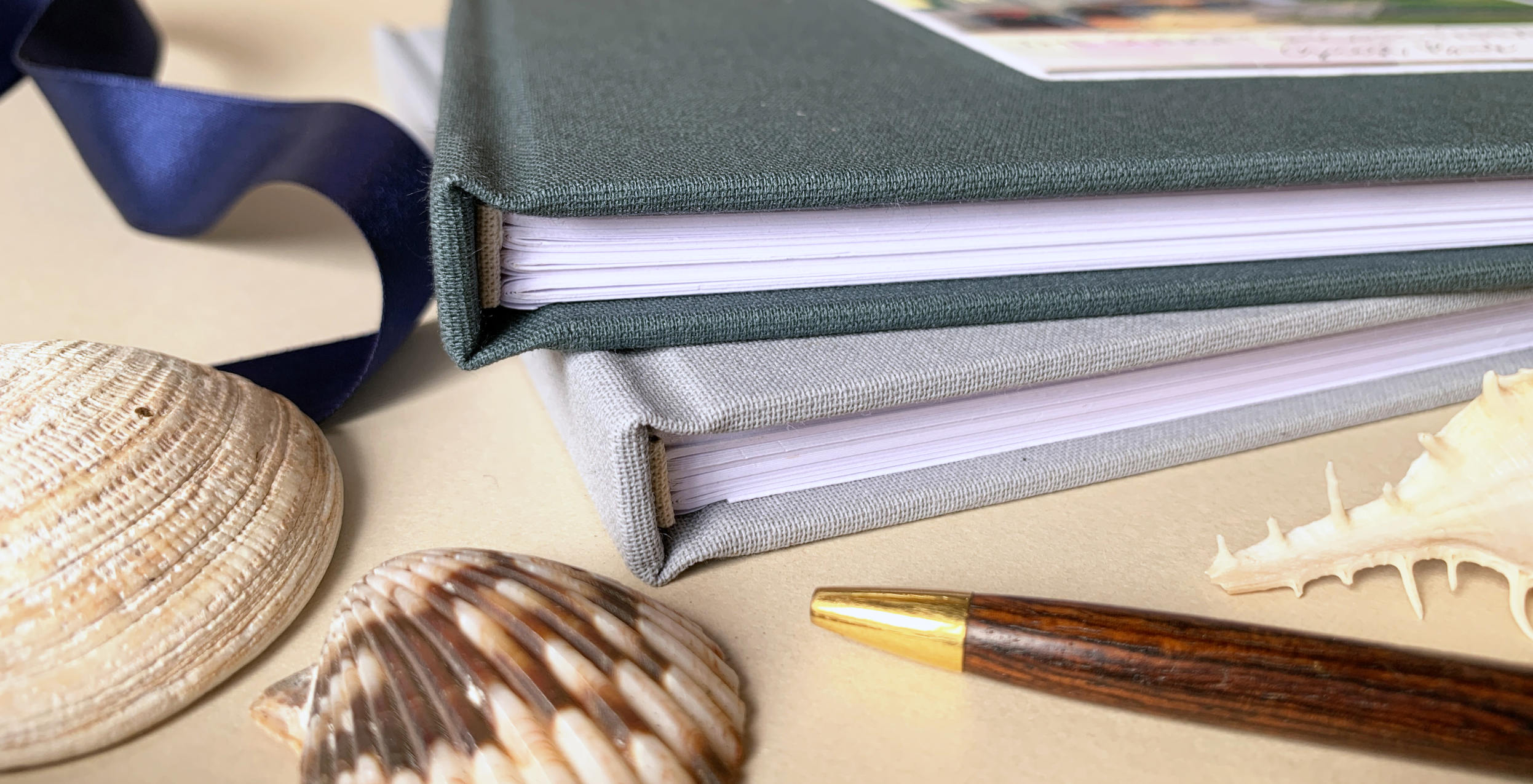 Close up photo of personalized linen guest books in dark and light gray with sea shells, ribbon and a pen.
