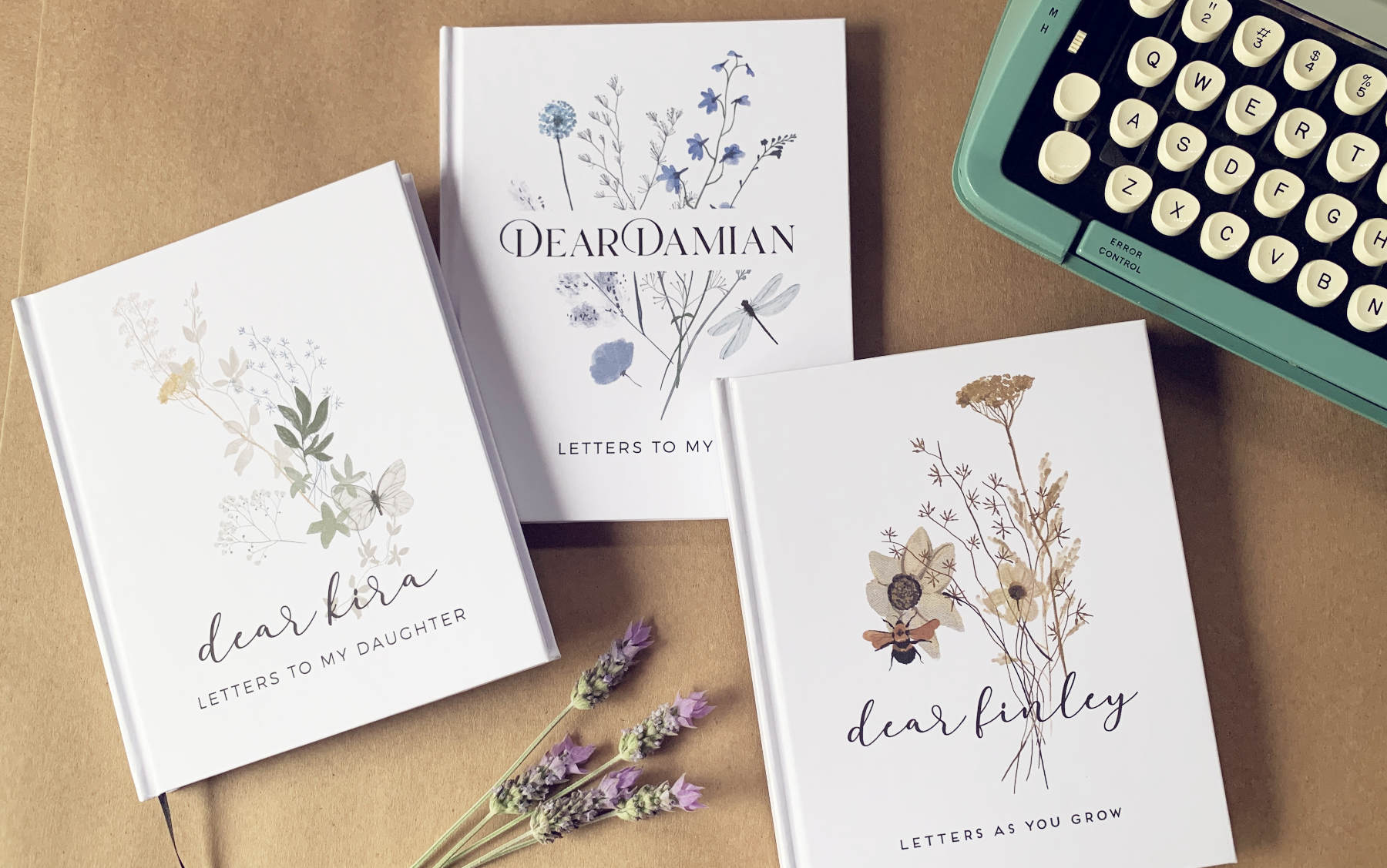3 personalized pregnancy journals on a table next to a typewriter and lavender flowers. each cover has a different wildflower design and custom names.