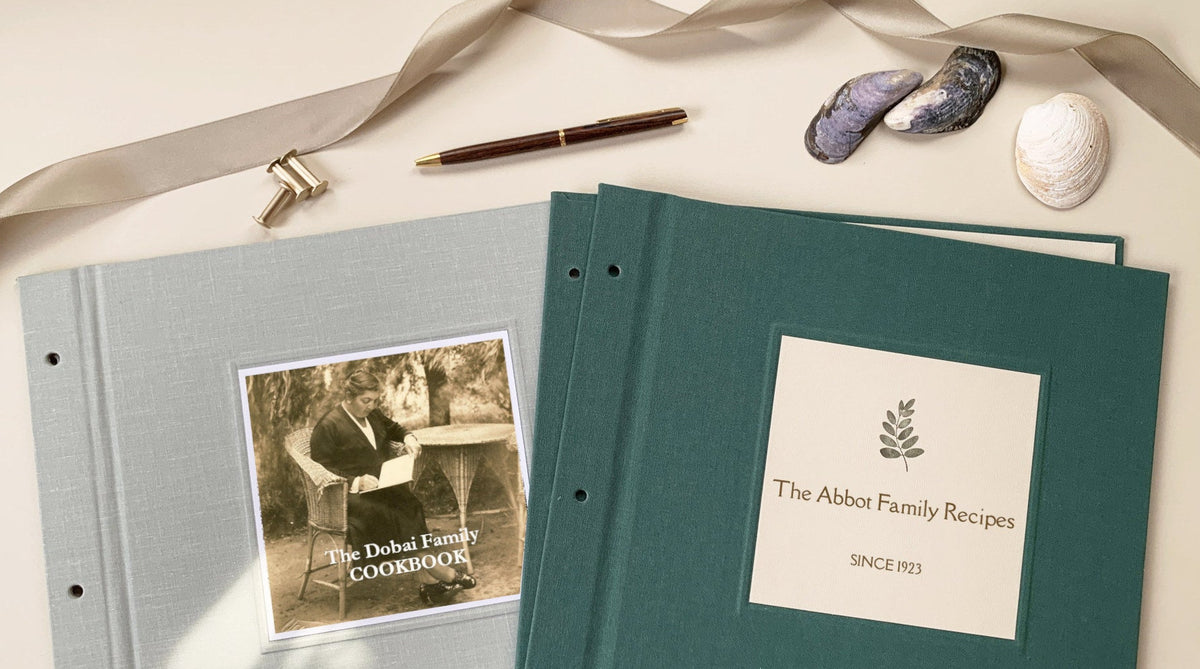 A gray linen and a forest green linen recipe binder displayed on a beige table with a ribbon, sea shells and a pen and brass screwposts. Each binder cover has a square inlay personalized with text and a photo (left) and a leaf (right).
