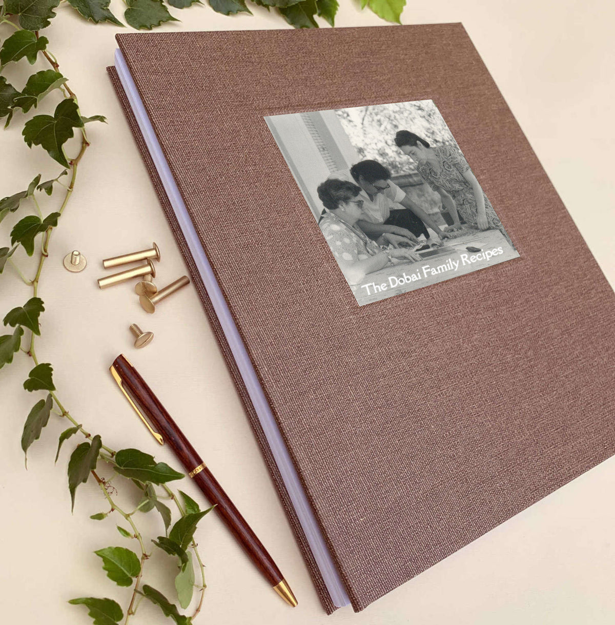 A personalized hardcover recipe book binder with a brown linen cover and a black-and-white family photo label, styled on a light beige background with green leaves, brass screwposts, and a pen.