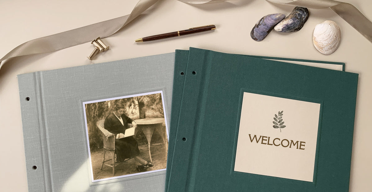 A gray linen scrapbook album next to a forest green scrapbook album displayed on a beige table top with brass screwposts, a pen, shells and a decorative ribbon.