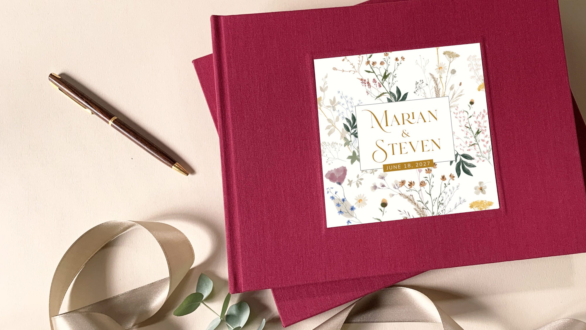 A personalized hardcover wedding guest book in deep magenta with a floral label reading 'Marian & Steven', styled on a cream background with a pen, ribbon, and eucalyptus leaves.