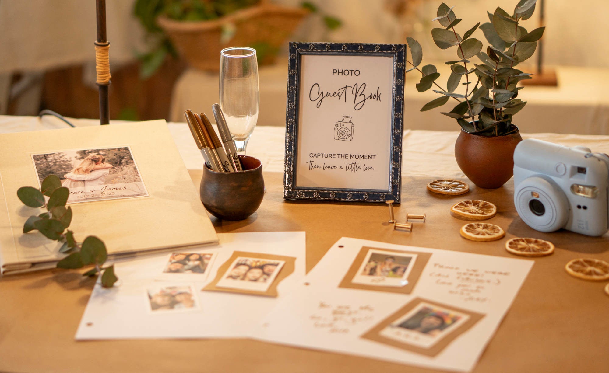 Party table setup with photo guest book, stationery, camera, table sign, pens, and decorative items on a wooden surface.